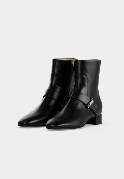 Paire de bottines en cuir noir à bouts pointus, fermeture éclair latérale, bride et talons blocs bas sur fond blanc.