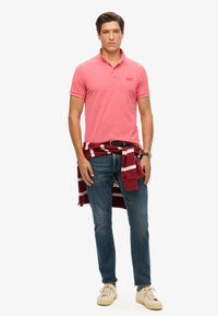 Camisa polo rosa, jeans de mezclilla y zapatillas beige claro. Sudadera a rayas en rojo oscuro y blanco atada a la cintura. Reloj plateado en la muñeca.