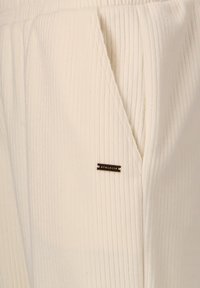 Weiße, gerippte Jogginghosen mit einer sichtbaren Tasche und einem kleinen schwarzen "ATHLETICA"-Logo-Tag, das an der Seite genäht ist. Glatte Textur.