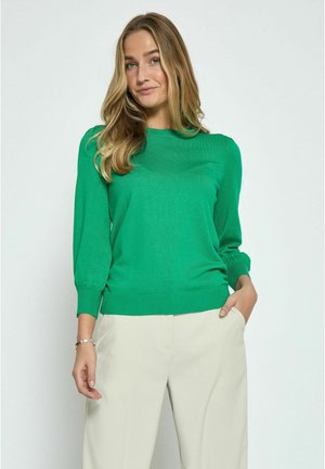 Groene gebreide trui met een ronde halslijn en aansluitende mouwen, gecombineerd met lichtbeige broek. Het materiaal lijkt zacht en glad.