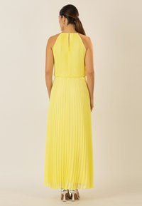 Gelbes, plissiertes Maxikleid mit Halterneck, Rückenschlitzdetail und fließendem Stoff, der bis zu den Knö cheln fällt. Sichtbare silberne High-Heels.