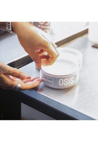OSiS+ Curl Jam stylar gel i en rund, vit burk med en grå etikett. En hand applicerar gel från burken. Rostfri stålyta.