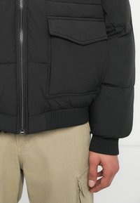 Schwarze gepolsterte Jacke mit Reißverschluss, gerippten Bündchen und einer Seitentasche. Verfügt über einen strukturierten Stoff und ein gestepptes Muster. Lockere Passform.