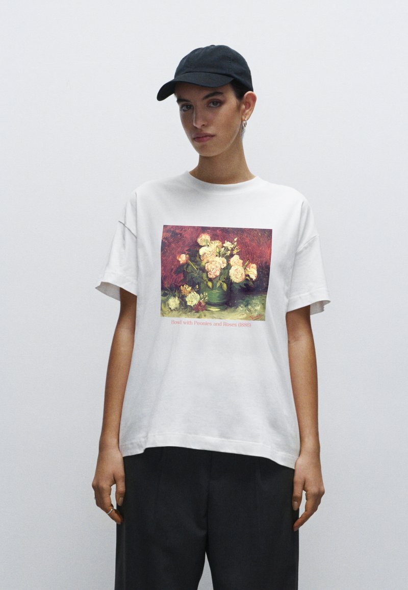Hvid bomulds T-shirt med et farverigt blomster maleri print. Sort kasket på, modellen står foran en ensfarvet hvid baggrund.