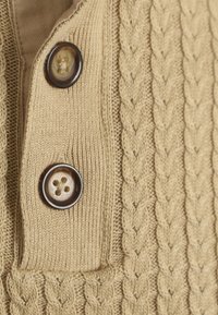 Beige gebreide trui met een kabelpatroon, een kraag en twee bruine knopen. De textuur is geribbeld en de stof oogt zacht.