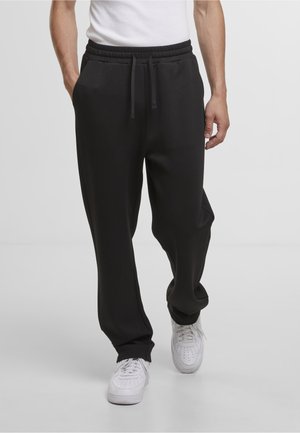 Homme portant un pantalon de survêtement noir avec taille élastique et cordon, assorti à des baskets blanches, marchant sur un fond clair uni.
