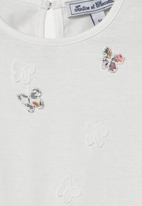 Valkoinen puuvillapaita, jossa on teksturoidut brodeeratut perhoset valkoisena ja kuvioitua kangasta, selkäpuolella vetoketju ja pyöreä pääntie.