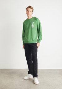 Grön sweatshirt med ribbade muddar och nederkant, prydd med ett vitt grafiskt emblem. Matcher med svarta byxor och vita sneakers på en ljus yta.