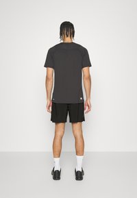 Camiseta negra de manga corta con cuello redondo y abertura lateral. Combinada con pantalones cortos negros. Modelo visto desde la parte posterior, de pie sobre un suelo blanco.