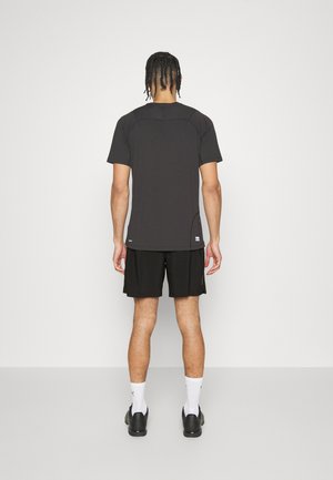 Camiseta negra de manga corta con cuello redondo y abertura lateral. Combinada con pantalones cortos negros. Modelo visto desde la parte posterior, de pie sobre un suelo blanco.