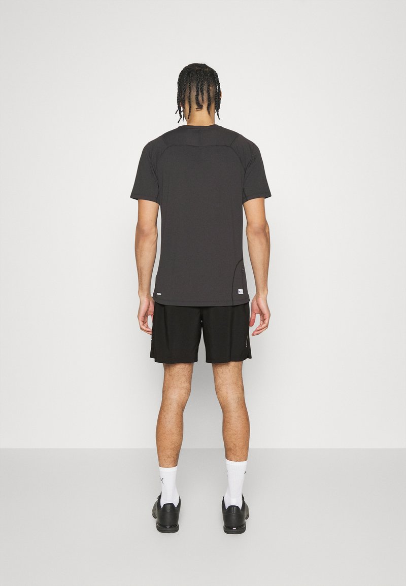 Camiseta negra de manga corta con cuello redondo y abertura lateral. Combinada con pantalones cortos negros. Modelo visto desde la parte posterior, de pie sobre un suelo blanco.