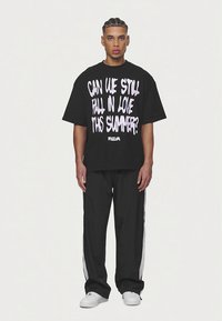 Μαύρο υπερμεγέθες μπλουζάκι με έντονο λευκό κείμενο: «ΜΠΟΡΟΥΜΕ ΑΚΟΜΑ ΝΑ ΕΡΩΤΕΥΤΟΥΜΕ ΑΥΤΟ ΤΟ ΚΑΛΟΚΑΙΡΙ;» Συνδυάζεται με μαύρο παντελόνι φόρμας με λευκές ρίγες.