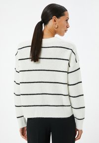 Pull blanc avec des rayures horizontales noires, présentant une coupe décontractée, une texture côtelée et des manches longues. Conçu avec un col rond.