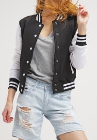 Svart och vit collegejacka med ribbad muddar, tryckknappar och kontrasterande ärmar, kombinerad med ljusblå slitna denimshorts.