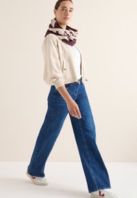 Giacca beige corta, t-shirt bianca, jeans a gamba larga blu e sciarpa color bordeaux con motivo. Le calzature presentano suole chiare con dettagli colorati.