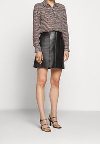 Vrouw in een gestructureerde blouse met lange mouwen, een zwarte leren minirok met een ritssluiting aan de voorkant, en zwarte gestrikte hoge hakken, staande.