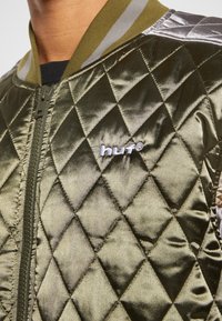 Gros plan sur une personne portant une veste matelassée en satin vert olive avec une fermeture éclair devant et le logo "huf" sur la poitrine.