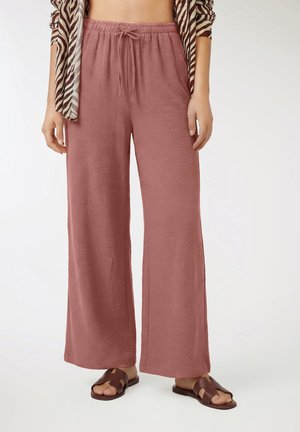 Kobieta ubrana w luźne, wrzosowe spodnie ze ściągaczem o teksturowanej tkaninie, zestawione z brązowymi sandałami typu klapki i bluzką w wzór zebry.