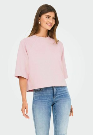 Vrouw met lang bruin haar glimlacht, gekleed in een lichtroze losse top met korte mouwen en lichtblauwe aansluitende jeans tegen een effen achtergrond.