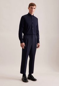 Camicia blu navy a maniche lunghe con bottoni, abbinata a pantaloni plissettati. Entrambi i capi presentano una texture liscia e una vestibilità su misura. Scarpe eleganti nere.