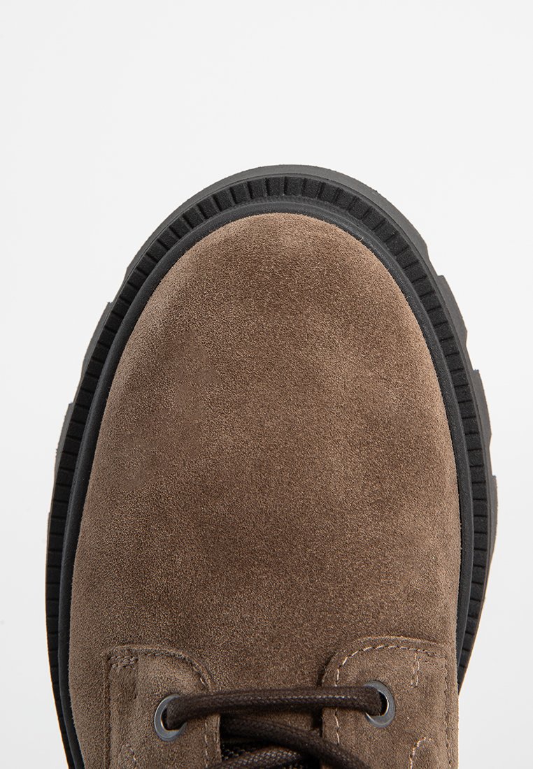Chaussure en daim marron avec bout rond, semelle en caoutchouc noir et lacets marron. Présente des détails de couture près des œillets et une surface texturée.