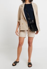 Žena stojí v béžovém saku přes námořnické triko, béžové kraťasy a černé sandály se cvočky, a drží malou černou crossbody kabelku.