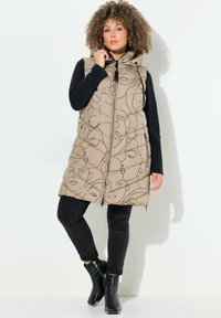 Beige gesteppte Weste mit abstrakten Gesichts-Linienmustern, einem Reißverschluss vorne und Kapuze, kombiniert mit schwarzen Slim-Fit-Hosen und Ankle-Boots.