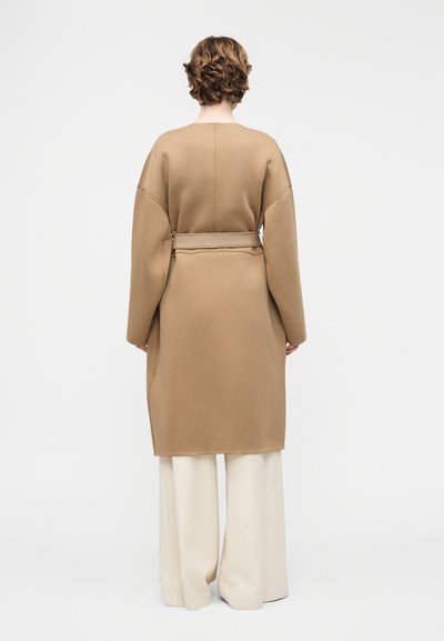 Άτομο με κοντά, σγουρά μαλλιά που φοράει ένα μακρύ παλτό σε χρώμα καμηλό με ζώνη και φαρδιά λευκά παντελόνια, στεκόμενο με την πλάτη προς τα έξω σε ένα απλό φόντο.