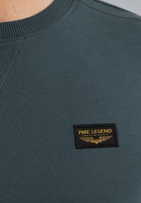Tamnozeleni dukserica s okruglim izrezom, koja ima teksturiranu crnu oznaku na kojoj piše "PME LEGEND AMERICAN CLASSIC" uz grafiku krila.