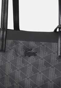 Sac cabas noir en tissu texturé présentant un motif géométrique, avec des anses synthétiques noires et un accent logo proéminent.