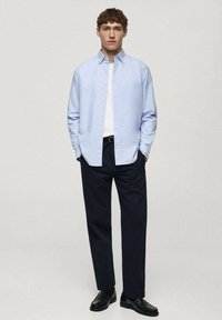 Chemise bleu clair à manches longues avec col, portée par-dessus un t-shirt blanc, assortie à un pantalon sombre et des chaussures noires, se tenant devant un fond neutre.
