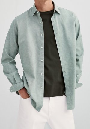 Homme portant une chemise vert clair à boutons ouverte sur un t-shirt vert foncé avec un pantalon blanc, mains dans les poches, sur fond uni.