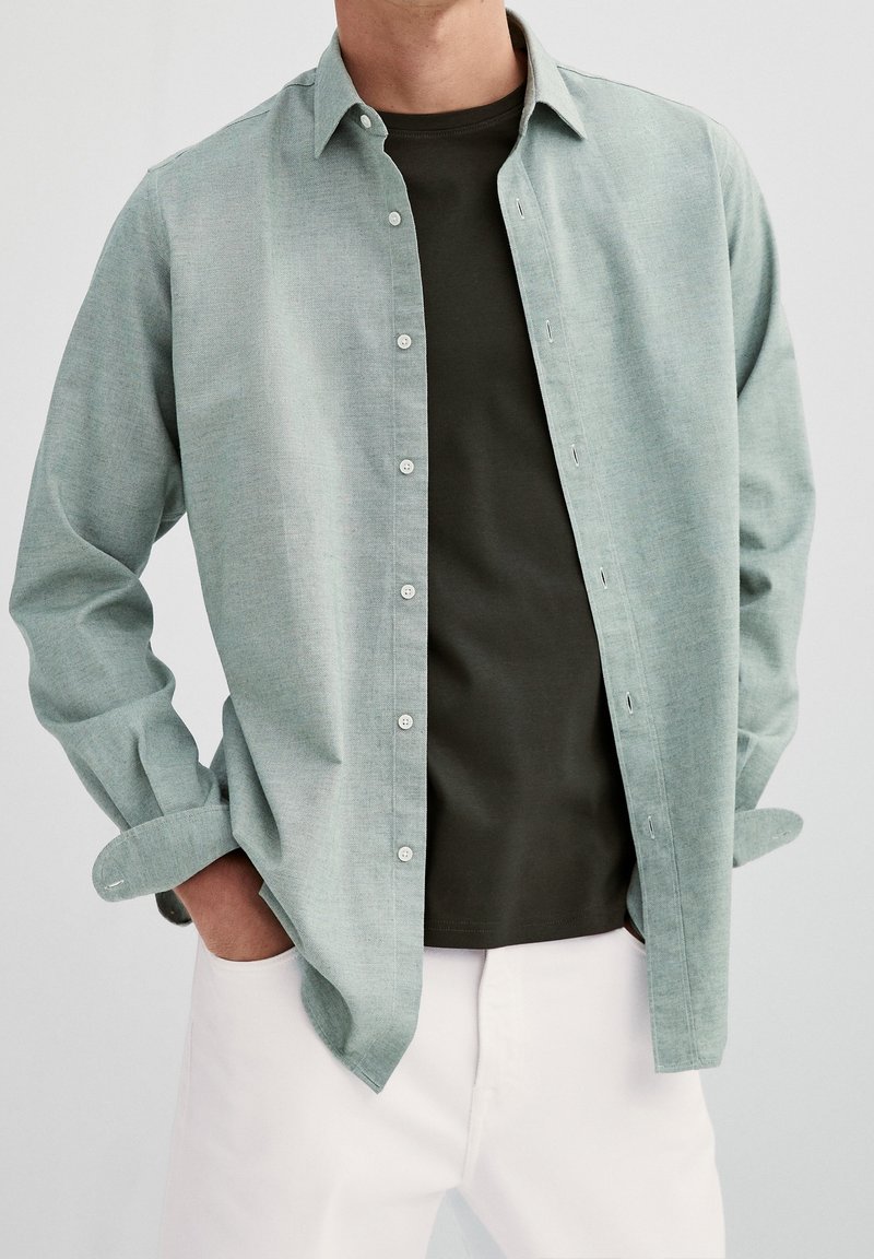 Homme portant une chemise vert clair à boutons ouverte sur un t-shirt vert foncé avec un pantalon blanc, mains dans les poches, sur fond uni.