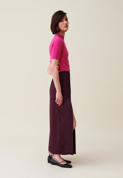 Maglietta aderente rosa a maglia con maniche corte, abbinata a una lunga gonna viola scura con uno spacco laterale. Completano il look delle scarpe basse nere.