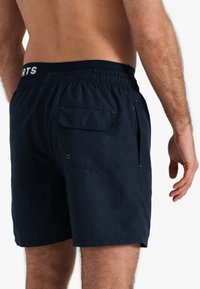 Marineblauwe zwembroek met een elastische tailleband, zijzakken en een achterzak met een knoopsluiting. Glad textuur.