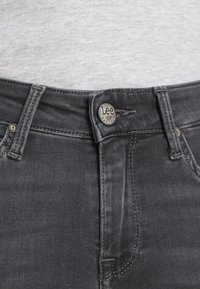 Jeans de mezclilla gris oscuro con cierre de botón de metal. Presentan costuras en contraste y un diseño estándar de cinco bolsillos. La textura parece suave.
