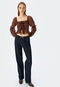 Braunes, geschopftes Crop-Top mit transparenten Puffärmeln, kombiniert mit dunkelblauen, weit geschnittenen Jeans. Das Outfit zeichnet sich durch ein figurbetontes Design und einen lässigen Stil aus.