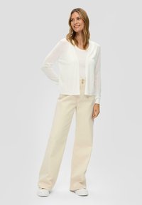 Witte cardigan over een licht tanktop, gecombineerd met beige wijde broeken. De outfit heeft een ontspannen pasvorm en een eenvoudig ontwerp, aangevuld met witte sneakers.