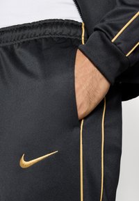 Čierne športové nohavice s zlatým logom Nike, obsahujúce bočnú kapsu a kontrastné zlaté akcentové pruhy pozdĺž švov. Mäkka textúra.