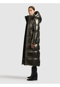 Sieviete stāv sāniski, valkājot garu metālisku pufojošu mēteļa ar kapuci un melnas slēptās platformas zābakus pret vienkāršu pelēku fonu.