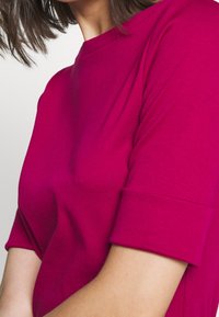 Fuksia T-paita pehmeästä kankaasta, jossa on pyöreä kaula-aukko, käärityt lyhyet hihat ja rento istuvuus, korostaen sileää rakennetta.