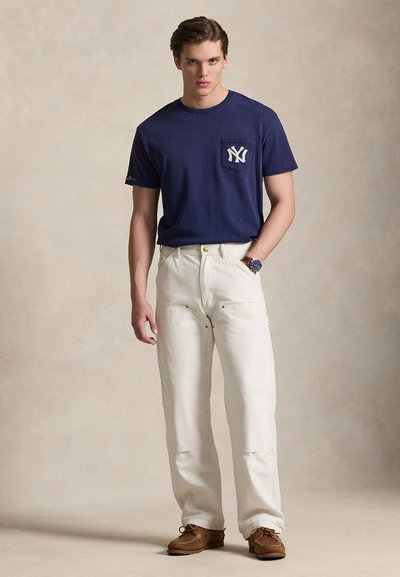 T-shirt en coton bleu marine avec poche avant et logo NY blanc, associé à un pantalon blanc ample et des chaussures marron à lacets. Texture lisse.