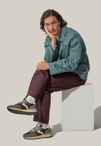 Modella che indossa un giaccone turchese chiaro, una camicia bianca, pantaloni borgogna e sneakers grigio-marroni con accenti bianchi e suola texture, seduta su un piedistallo.