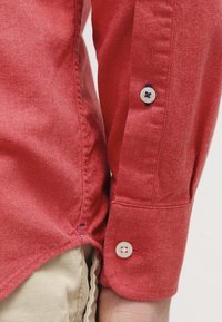 Rotes Button-up-Hemd aus strukturiertem Stoff, mit weißen Knöpfen und marineblauen Akzenten sowie einem abgerundeten Saum, teilweise gezeigt mit sichtbarem Manschette.