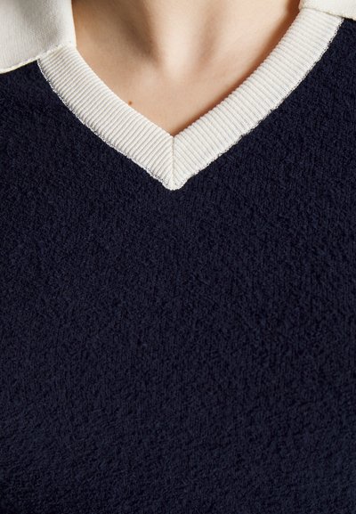 Tecido texturizado azul-marinho com gola em V; acabamento em branco canelado acentua o decote. O material parece suave e confortável.