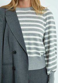 Siv l blazer s srajčno zapenjanjem. Spodaj je črtast pulover s krožnim izrezom v sivi in beli barvi. Mehak material, očitna je prilagojena moda.