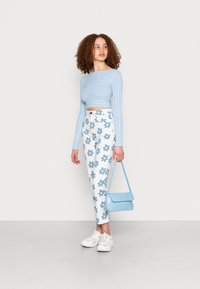 Suéter cropped canelado azul claro, calças brancas estampadas com flores, ténis brancos e uma pequena mala azul texturizada.