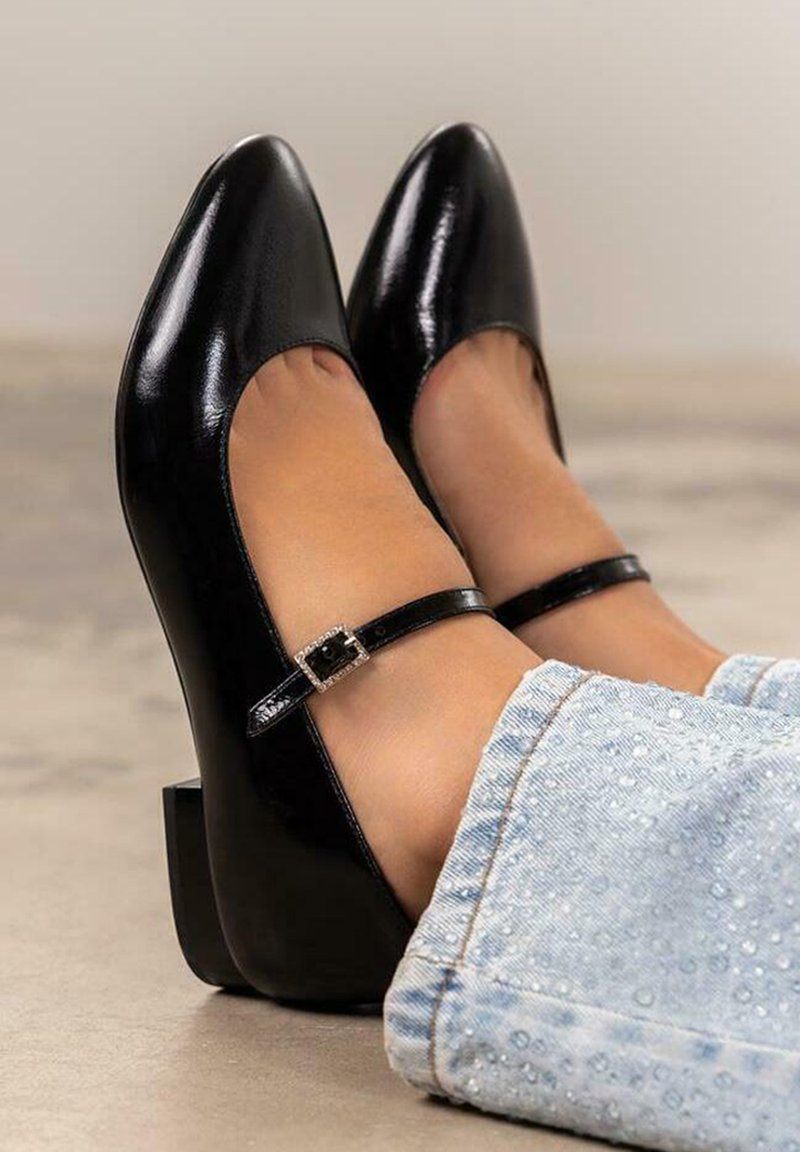 Zapatos de tacón de charol negro con punta afilada, tira al tobillo y tacón pequeño en bloque. Presentan un detalle decorativo de hebilla en la tira.