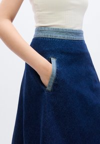 Τζιν φούστα με σκούρο μπλε σώμα και πιο ανοιχτή μπλε ζώνη μέσης, με καμπυλωτή τσέπη με άκρη από πιο ανοιχτό τζιν. Λείο υλικό, σχέδιο σε Α γραμμή.