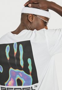 T-shirt blanche avec un grand motif de main en impression thermique colorée au dos sur fond noir. Manches courtes, en coton.
